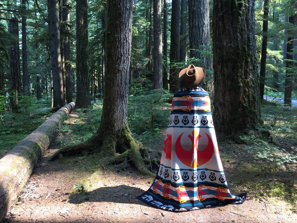 A woman stands at the edge of a forest wrapped in a Pendleton Star Wars blanket. 