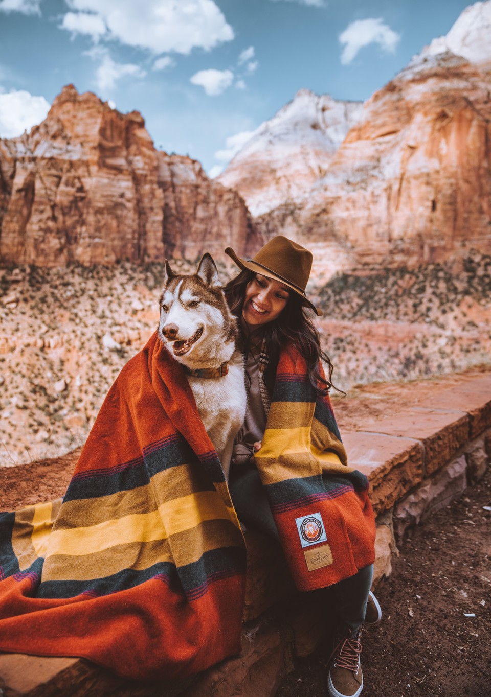 A woman wih a Husky dog sits in Zion National Park wrapped in a Zion blanket.