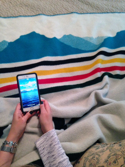 hands hold a cell phone showing a photo of Glacier National park, next to the silhouette of the glaciers on a Pendleton blanket. 