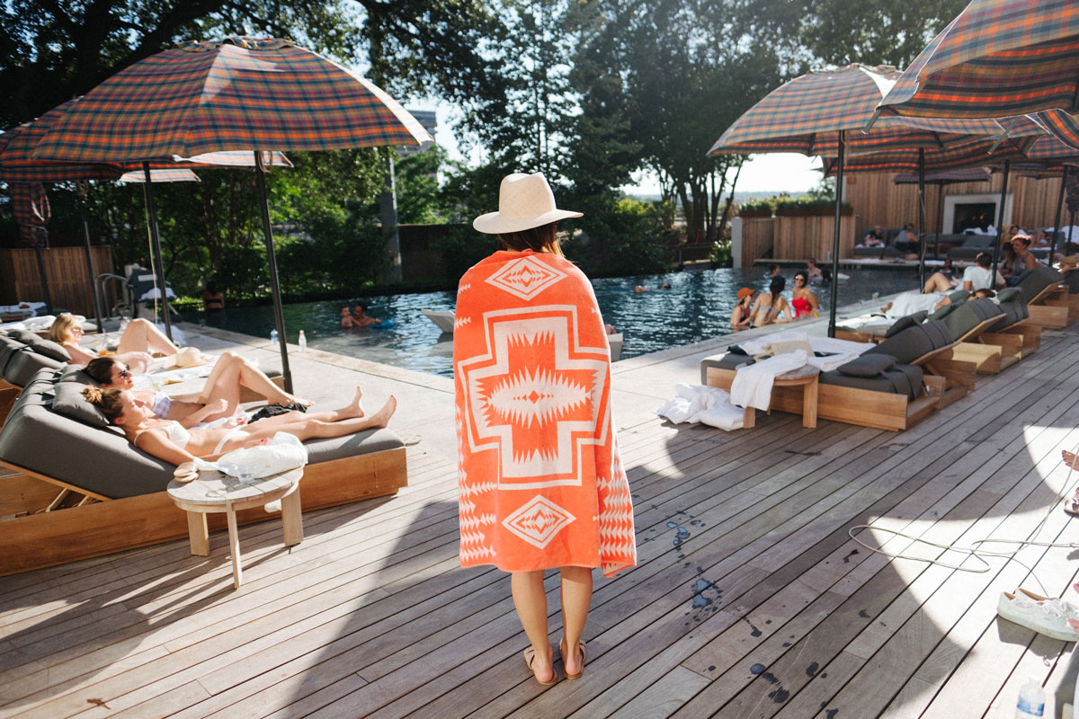 A young woman wrapped in an orange Pendleton beach towel stands on a wooden deck, looking at a swimming pool. Her back is to the camera and she is wearing a straw hat.