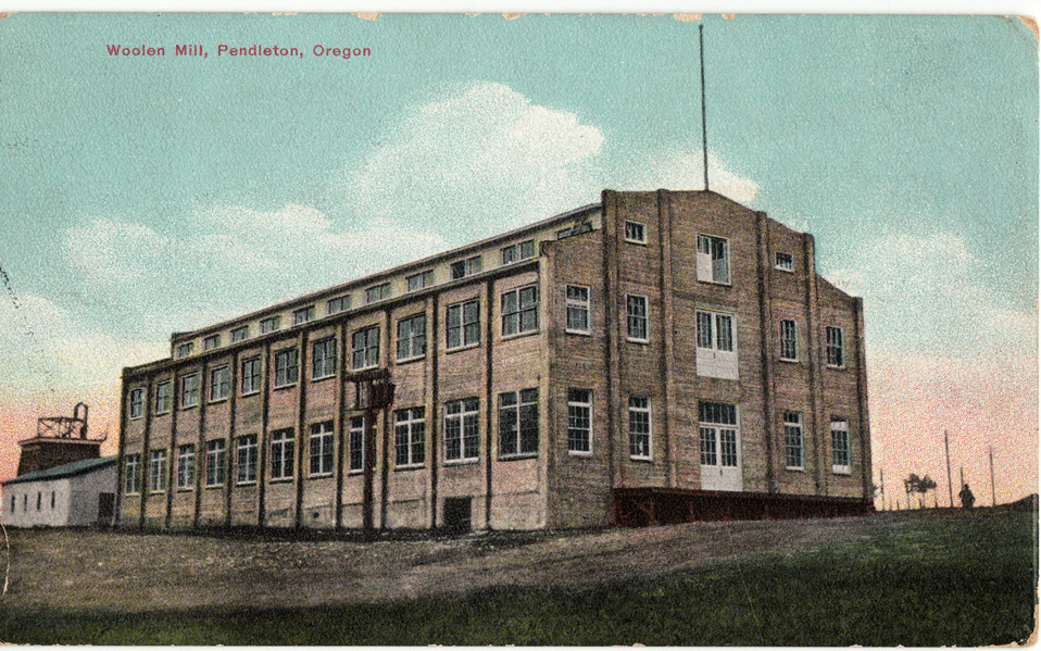 This photo is a vintage postcard image of the Pendleton, Oregon woolen mill. The building is grey brick, with rows of windows trimmed in white, and large front doors on the first, second and third floors at the front of the building.