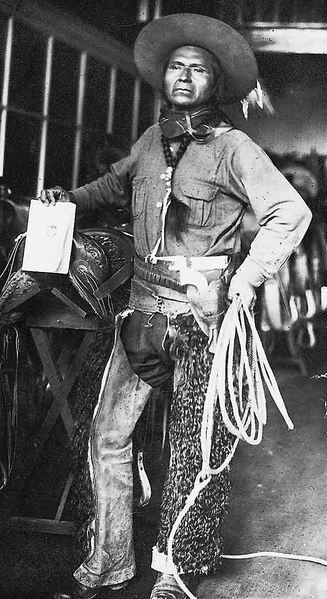 Jackson Sundown poses inside a stable with his championship saddle. He is wearing furry chaps, his signature hat, and a senim workshirt. When he rode, Sundown often tied his braids together on his chest, and they are so tied in this photo. He also holds a lariat.