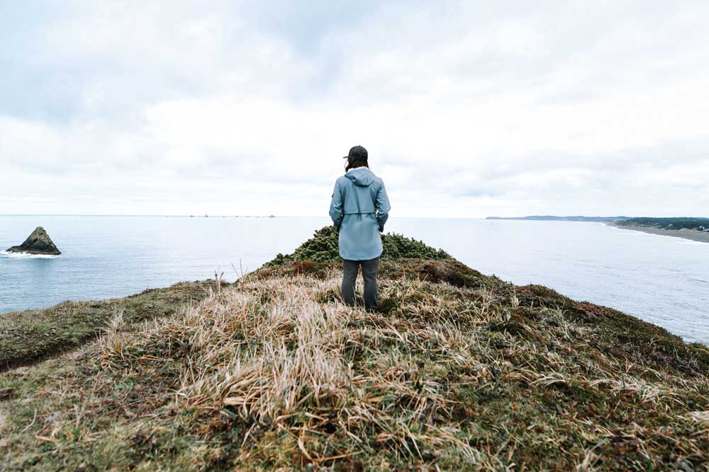 A woman stands on a grassy point, looking out over a bay. She is wearing a Pendleton rain slicker.