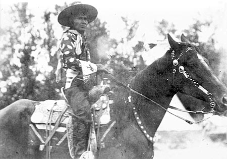 Jackson Sundown sits on his horse, wearing his signature hat, a dark cotton shirt with large light polka dots, and beaded gloves.