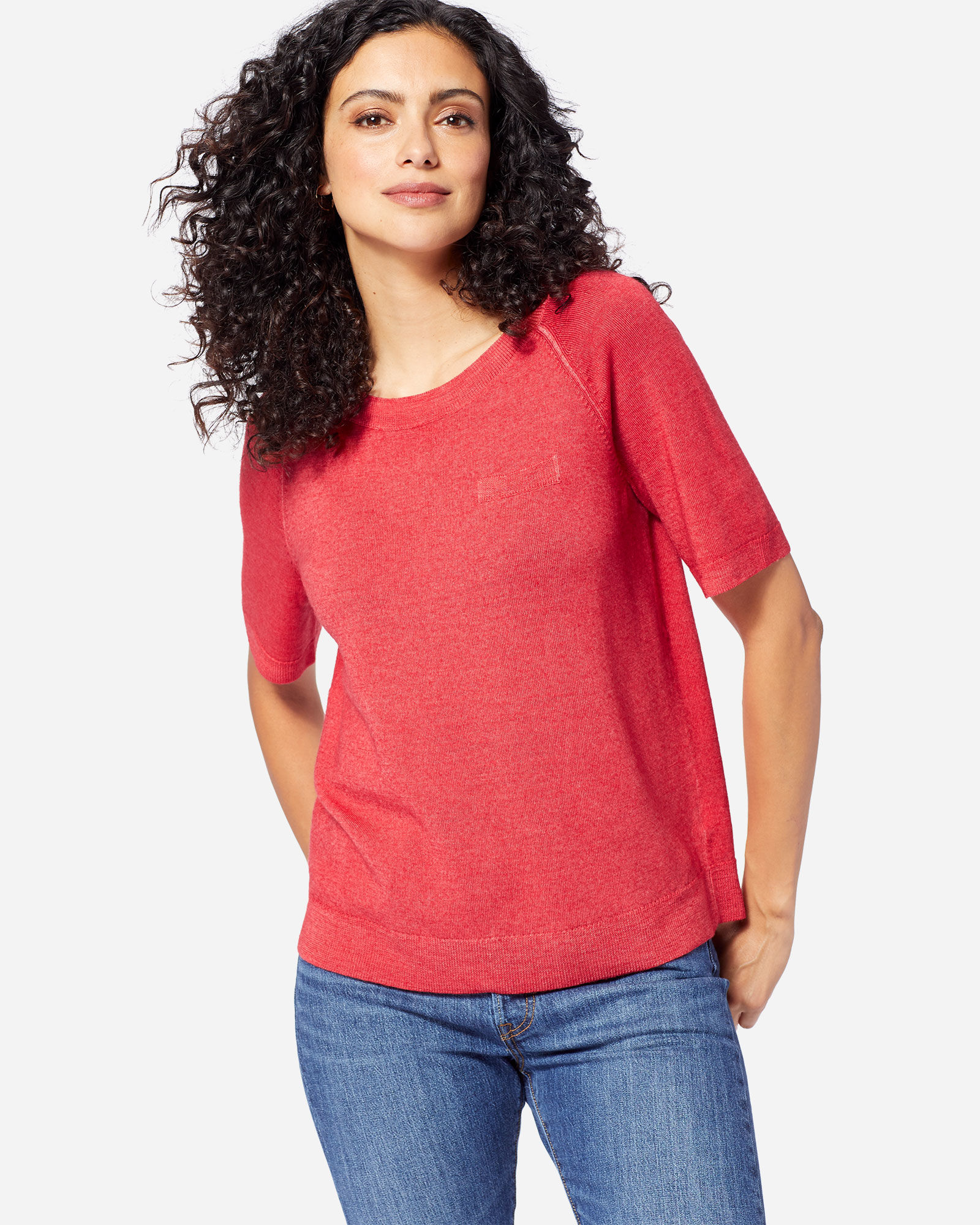 A woman with bountiful brunette curls stands with hands hooked in the back of her jeans pockets. She is wearing a brighter red sweater with short sleeves.