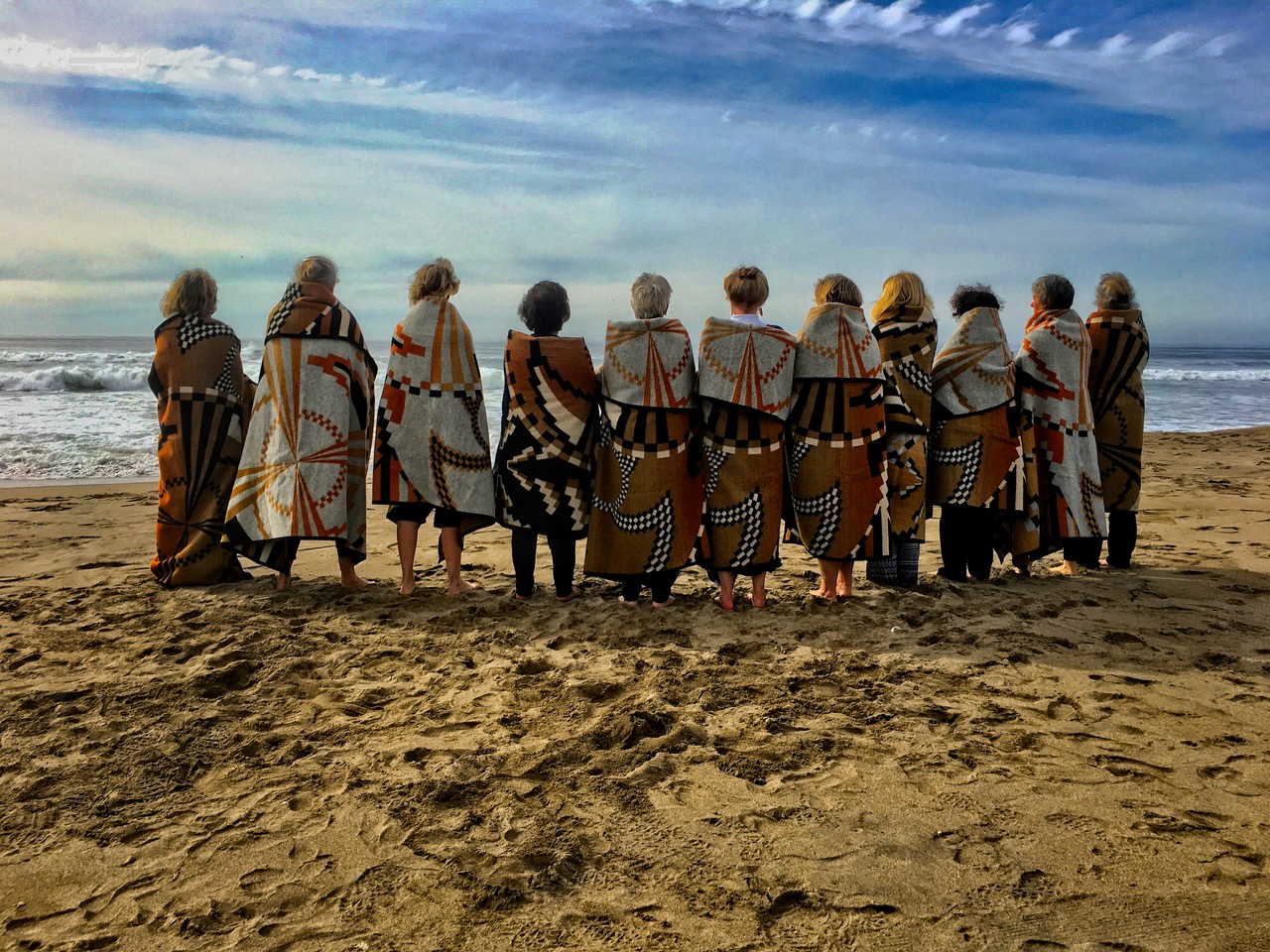 Final Ceremony: eleven women wrapped in Basket Dance blankets stand in a line at the beach.