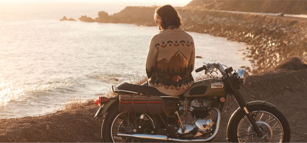 A man sits on a motorcycle overlooking the coastline. He is wearing a Pendleton sweater.