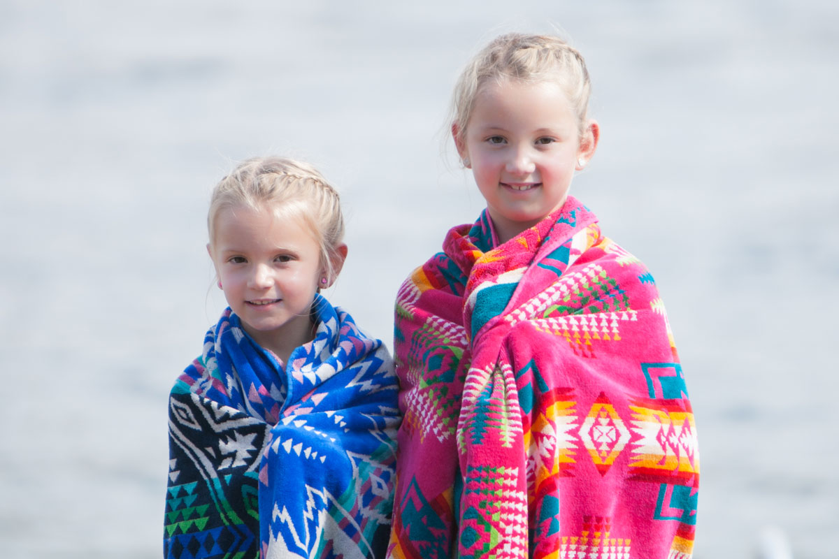 Two little girls who look like sisters, with blond braided hair, stand in front of water, wrapped in Pendleton beach towels.