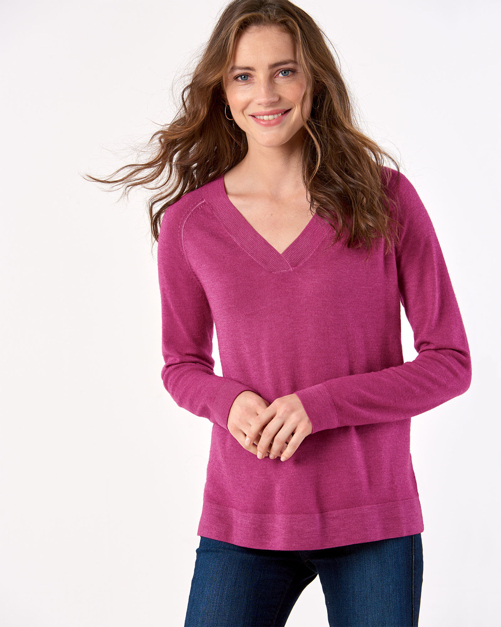 A woman with long brown hair and blue eyes stands before a white background, her hands held in front of her stomach. She is wearing a plum colored sweater and jeans.