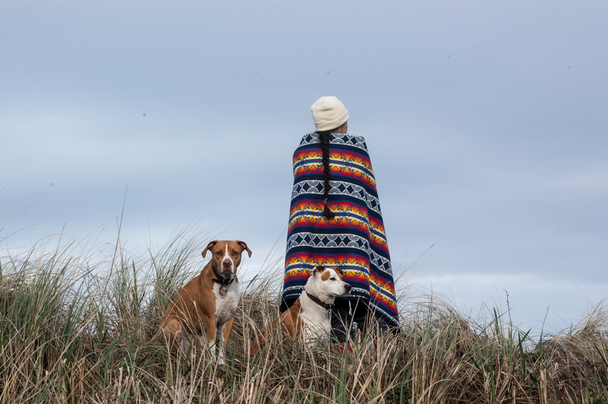 A woman shows off the We Walk Together blanket at Fort Stevens beach, along with two dogs.