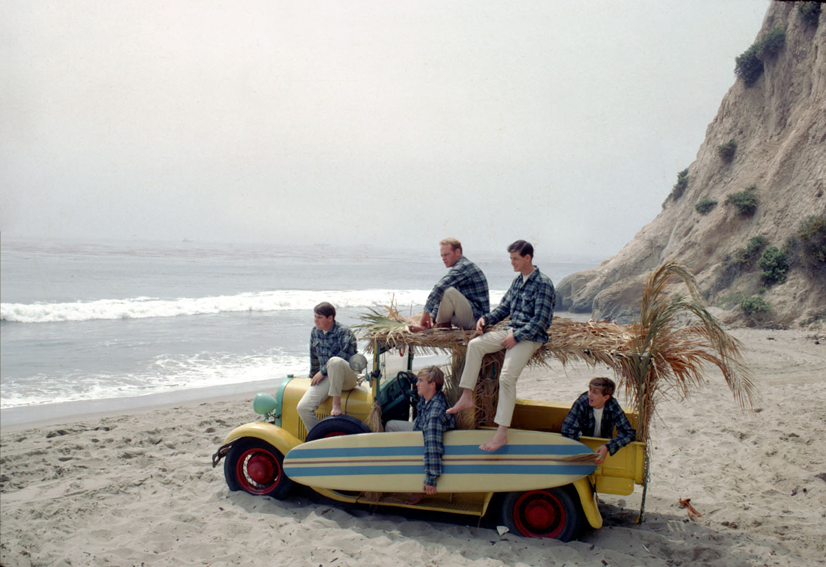 The Beach Boys in their Pendleton shirts, posing with a surfboard and palm-bedecked beach wagon on the California coast.