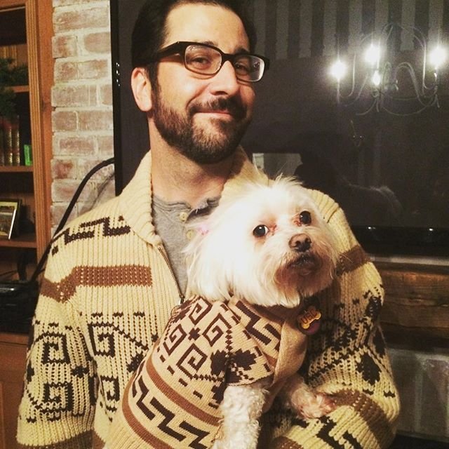 A man holds a white dog, they both are wearing Westerley sweaters