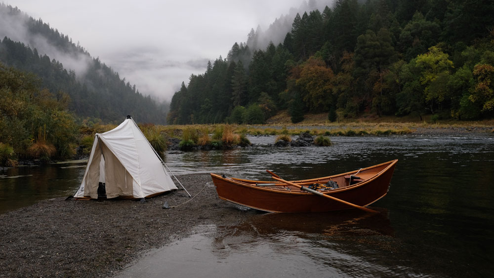 Morning on the river: a canvas tent and an empty wooden drift boat on a river at sunrise.