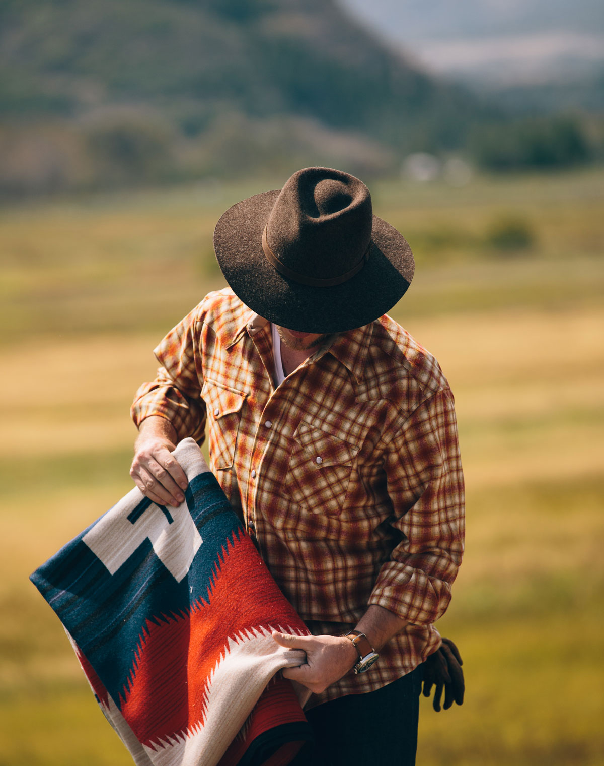 A man stands in a field before a mountain. He is wearing a plaid Pendleton shirt, and holding a folded Pendleton Brave Star blanket.