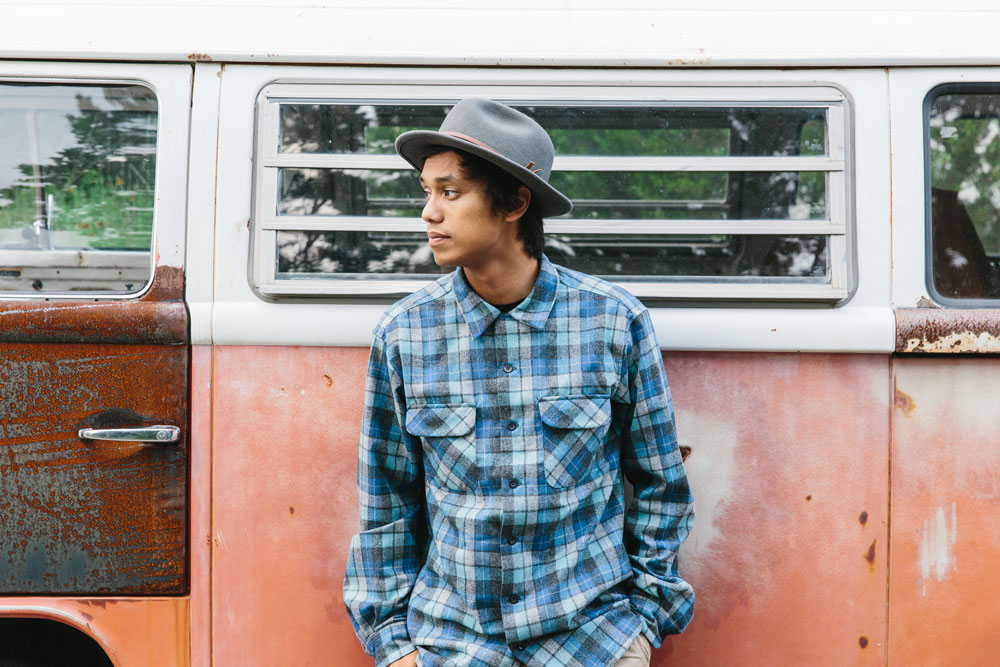 A young man leans against a rusted orange camper. he is wearing a Pendleton Board Shirt in Original Surf Plaid.