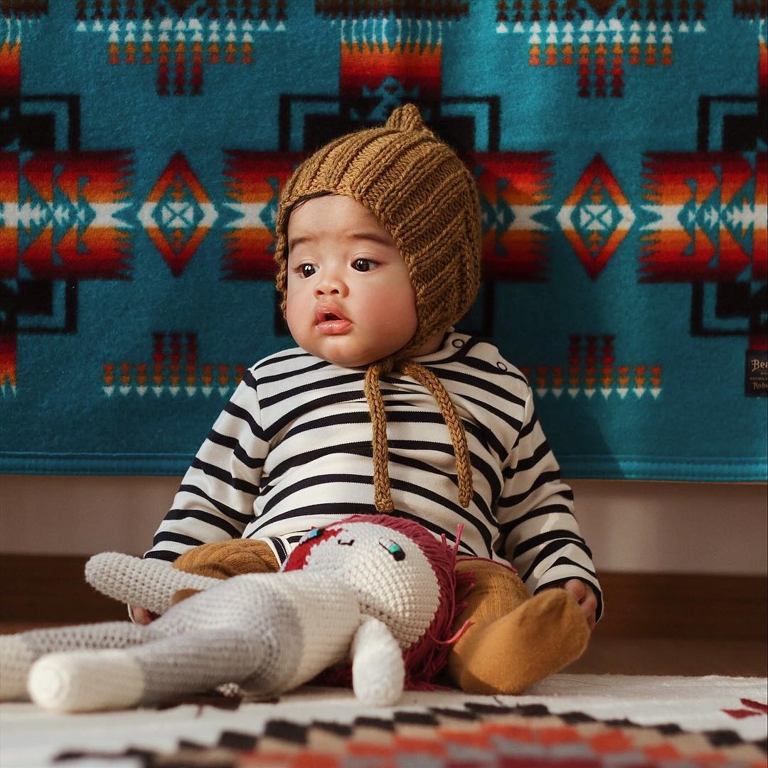 A sweet, chubby baby sits on a rug, leaning against a Pendleton baby blanket which is on a wall. The baby wears a kitted hat that ties under his chin, a striped shirt, and long knitted socks. A crocheted doll is on his lap. 