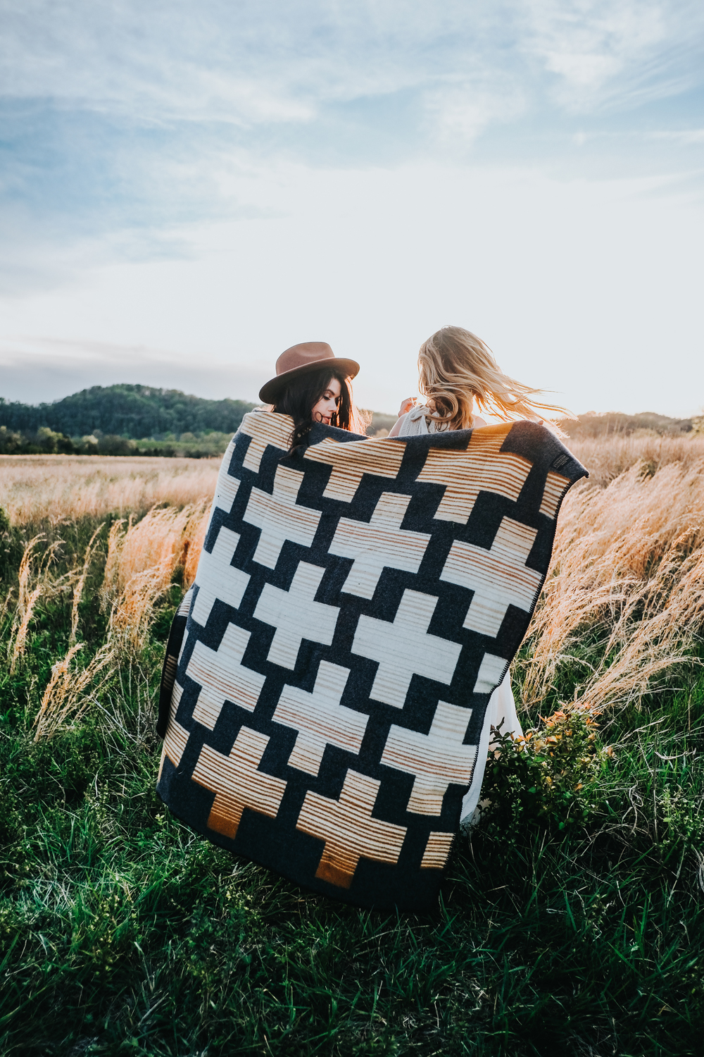 Two women wrap up in a blanket in a meadow.