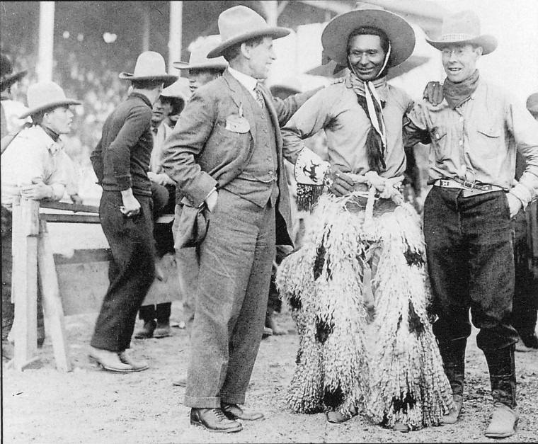 Jackson Sundown poses with another rider to his left and a businessman to his right. Sundown is smiling, and wearing his signature hat and silk kerchiefs, his wooly potted chaps, and beaded gloves that feature a rider on a bucking horse on the cuffs.