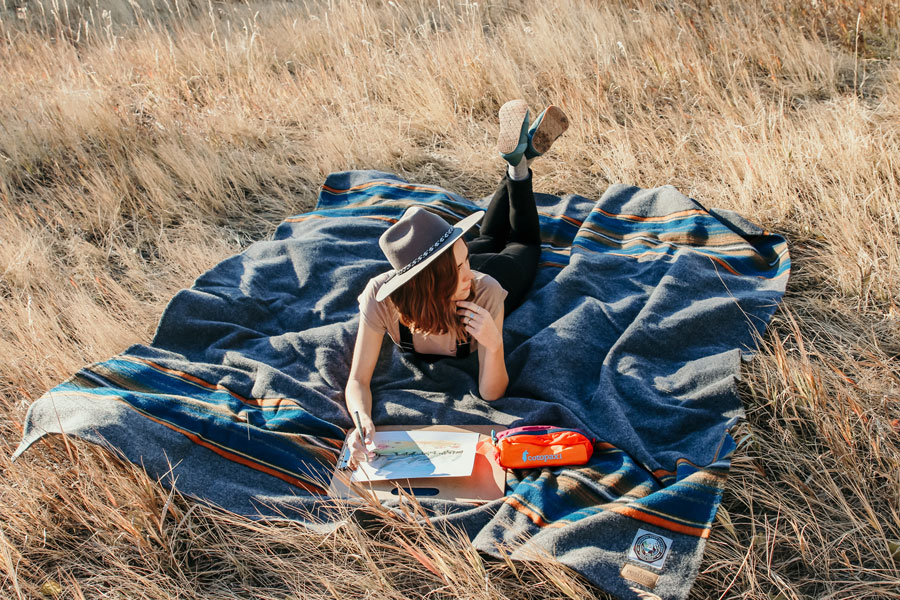 A young woman lays on her stomach on a Pendleton Olympic National park blanket, in a field of tall, dry grass. She is painting a watercolor.