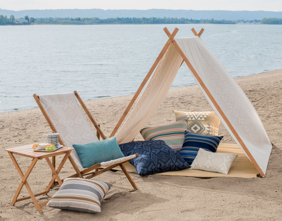 A sandy beach, with a folding chair, seven throw pillows, and tent made of Pendleton by Sunbrella fabrics.