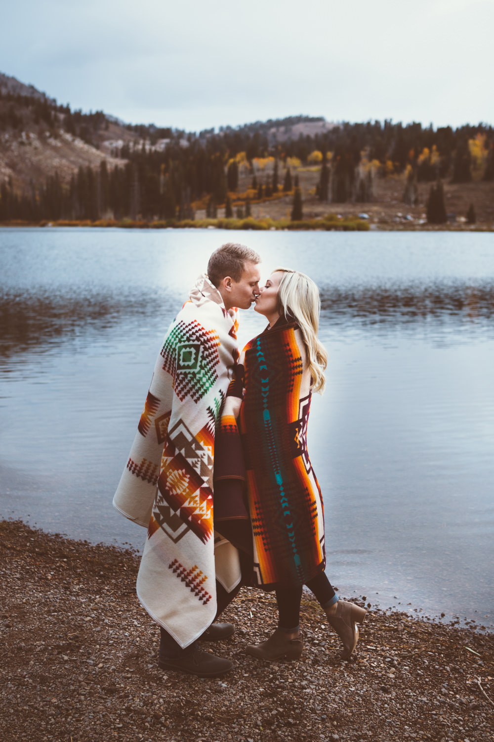 A couple kisses by a lake, wrapped in blankets. 